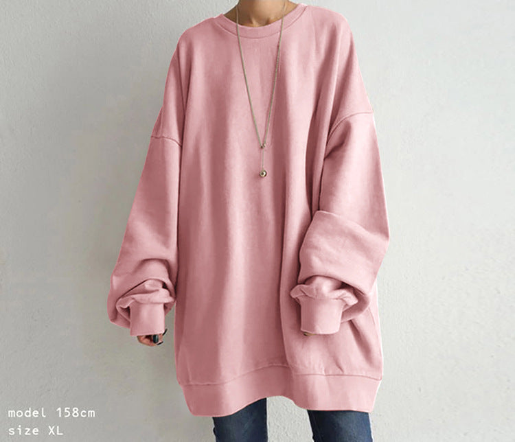 A person wearing an oversized dusty pink crewneck sweatshirt with cuffed long sleeves, paired with dark blue jeans and a long silver necklace, standing against a light gray wall.