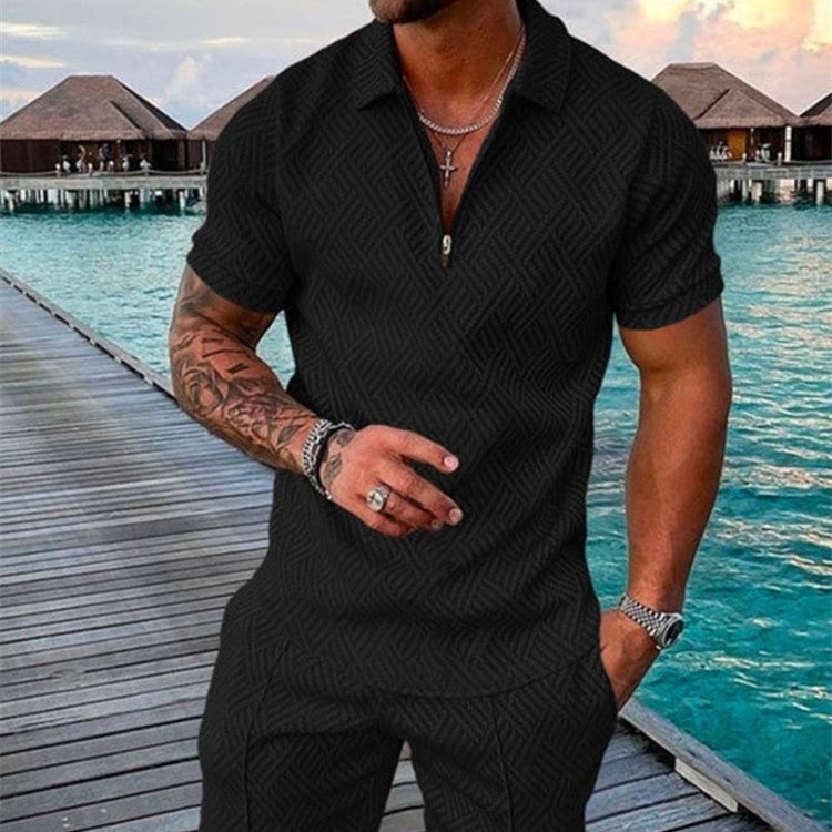 Man wearing a black textured polo shirt and pants by a waterfront with huts in the background.
