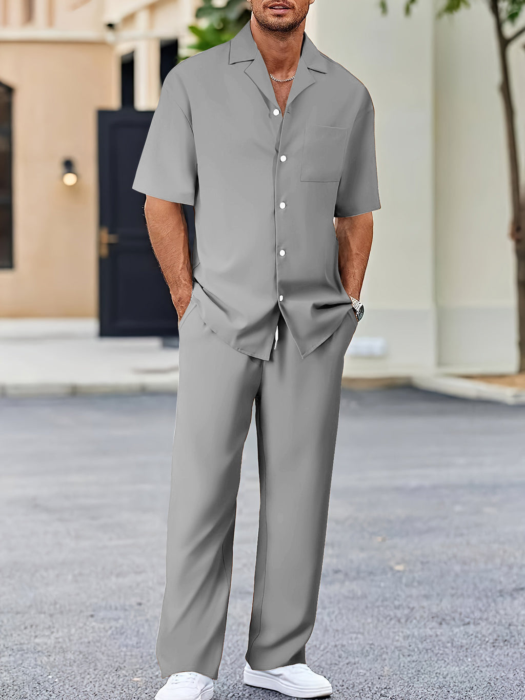 Man wearing a gray outfit standing outdoors with a building and tree in the background.