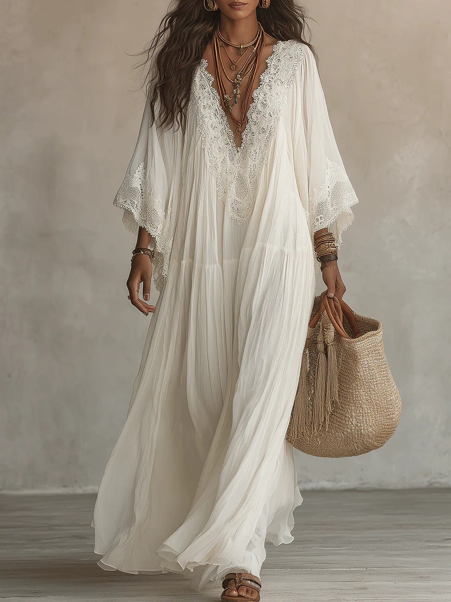 A woman wearing a flowing white bohemian maxi dress with lace trim and wide sleeves, holding a woven straw tote bag against a neutral textured wall.
