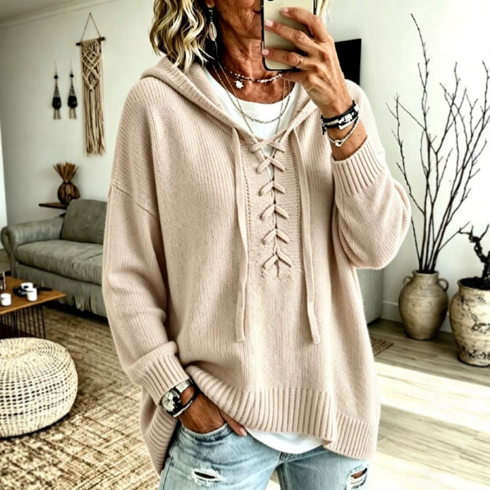 A woman takes a mirror selfie in a cozy living room, wearing a beige knit hooded sweater with lace-up front detailing, a white t-shirt, and distressed light blue jeans.