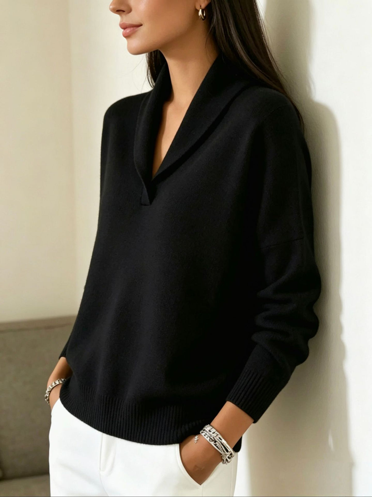 A woman wearing a black knit sweater with a wide shawl collar and white pants, leaning against a light-colored wall.