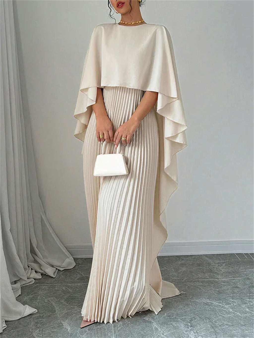 Woman wearing a beige cape over a pleated dress holding a white clutch against a plain background