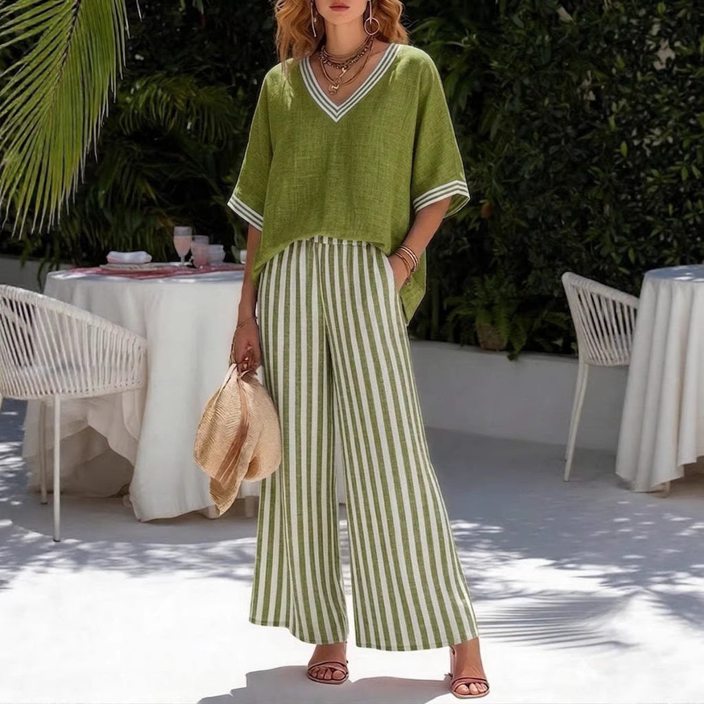 Woman in green top and striped pants standing outdoors with tables and chairs in the background.