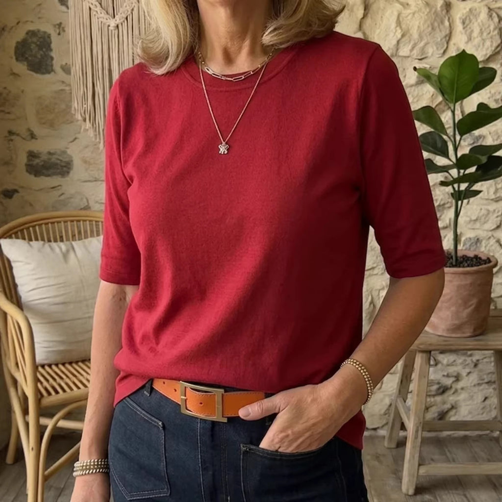 A woman wearing a red short-sleeve knit top tucked into dark blue jeans with a tan belt, standing in a rustic indoor setting with a stone wall, woven chair, and potted plant.