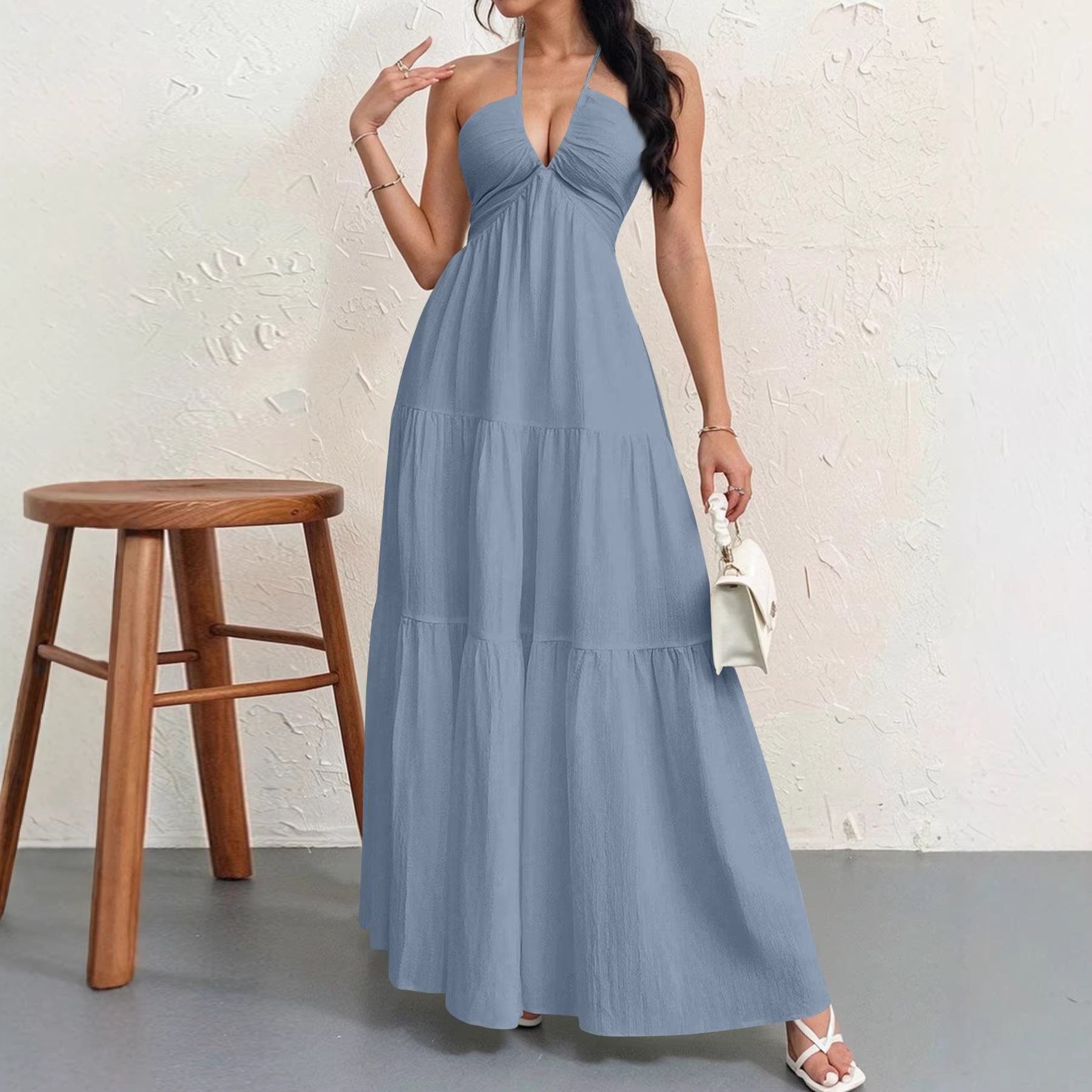 Woman wearing a long blue dress standing next to a wooden stool against a white wall.