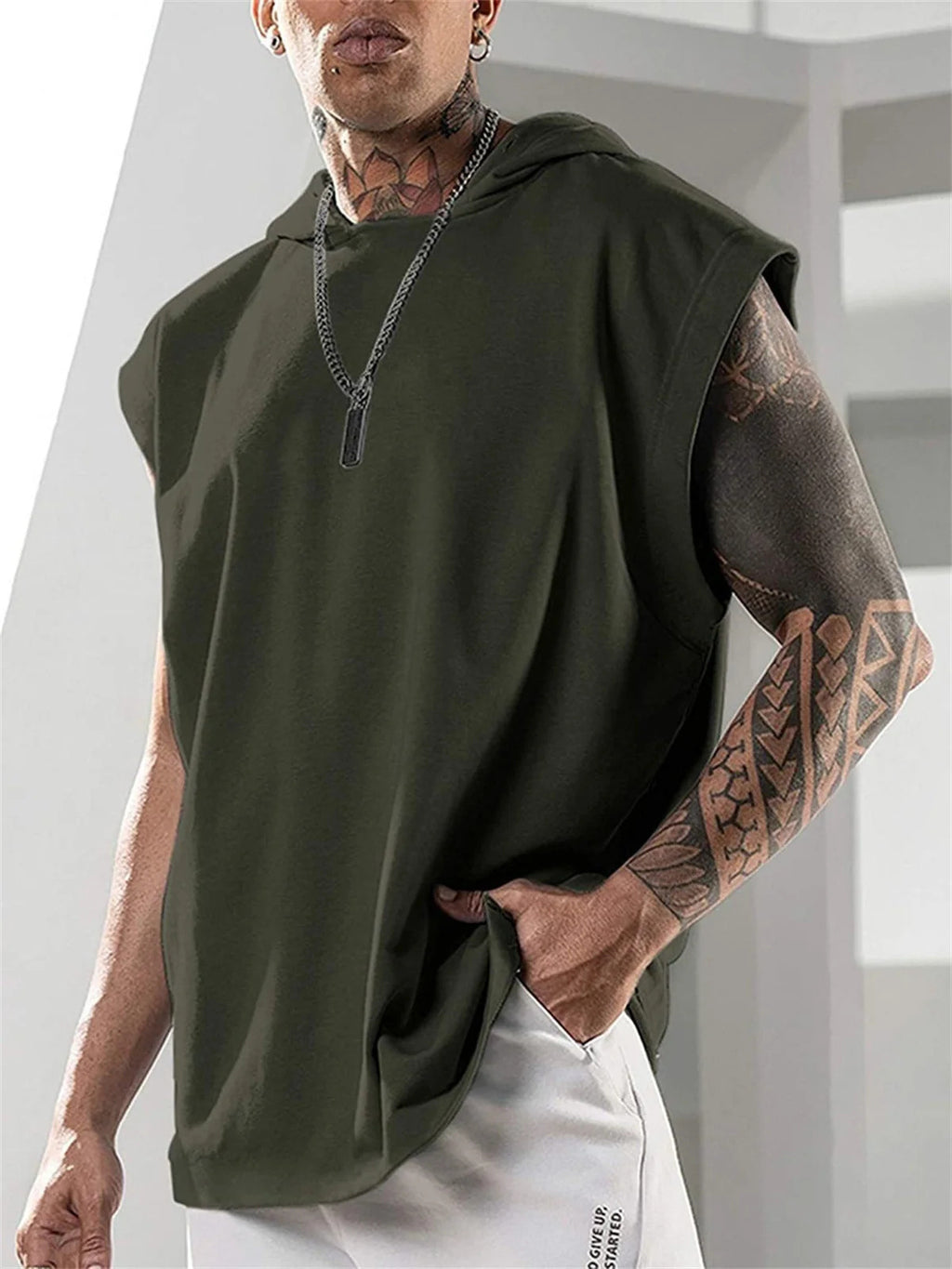 Person wearing a green sleeveless hoodie with tattoos on arms, standing indoors.