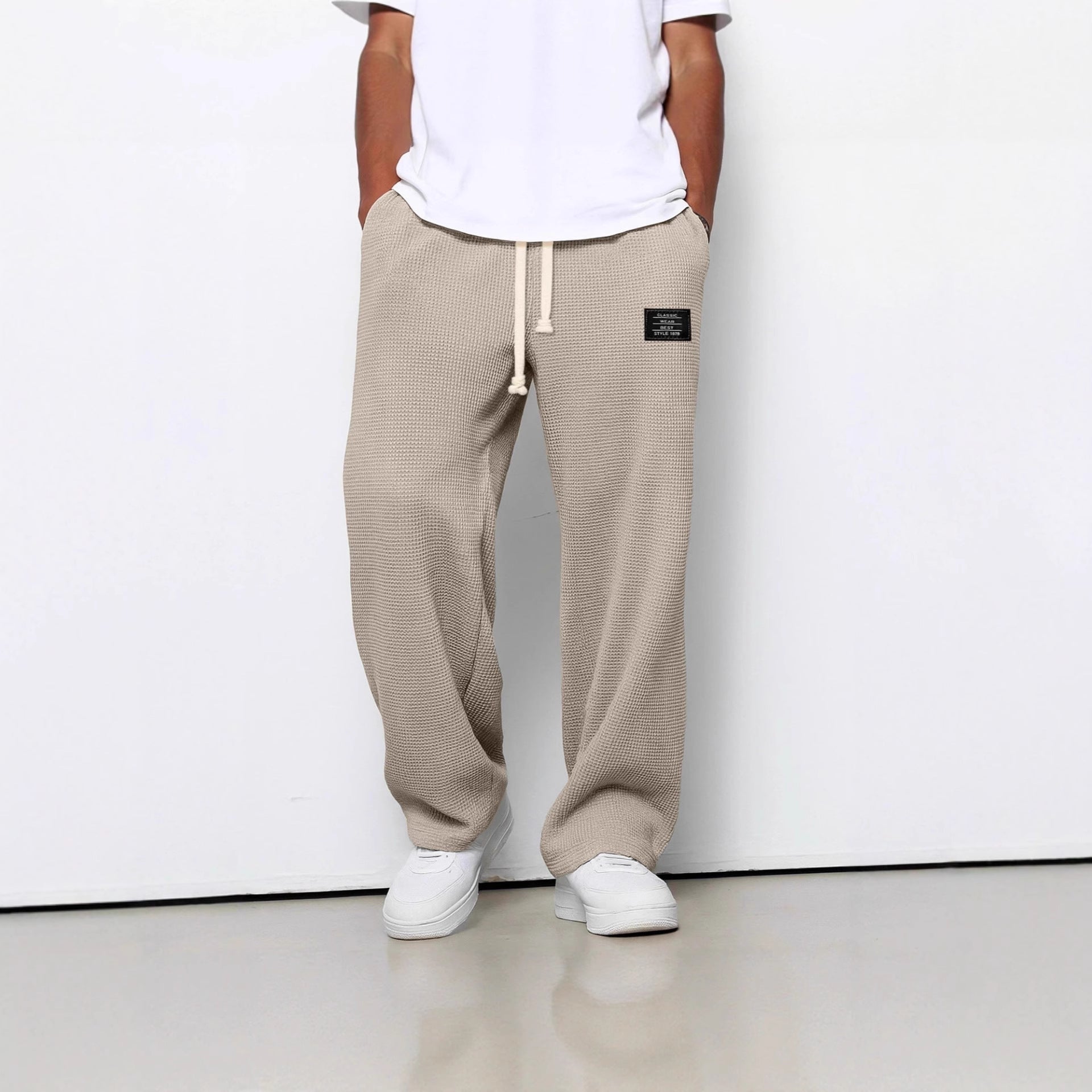 Person wearing beige sweatpants with a white shirt and white shoes on a plain background