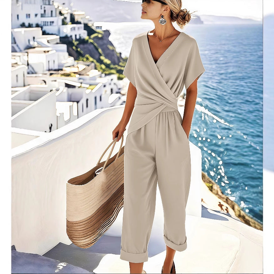 Woman in a beige jumpsuit standing on a balcony with a scenic view of a coastal town.