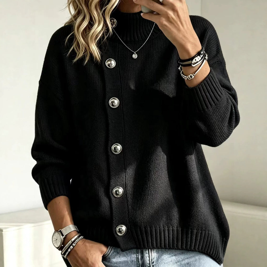 A woman wearing a black ribbed knit cardigan sweater with large silver buttons, paired with light blue jeans and silver jewelry, in a brightly lit indoor setting.