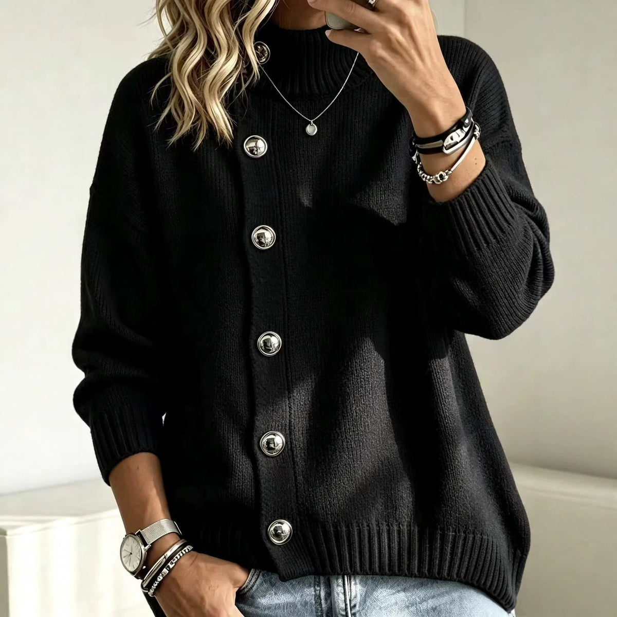 A woman wearing a black ribbed knit cardigan sweater with large silver buttons, paired with light blue jeans and silver jewelry, in a brightly lit indoor setting.