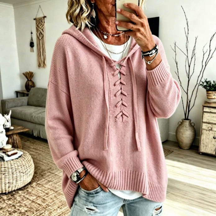 A woman takes a mirror selfie wearing a pink knit hooded sweater with lace-up front details, layered over a white top and paired with light blue distressed jeans in a cozy living room setting.