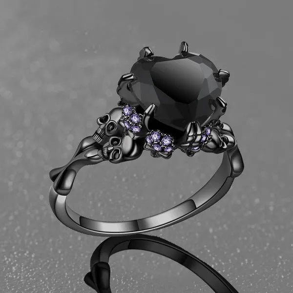 A gothic-style ring featuring a large faceted black heart-shaped gemstone, flanked by detailed skull and claw motifs set with small purple stones, shown on a textured gray background.