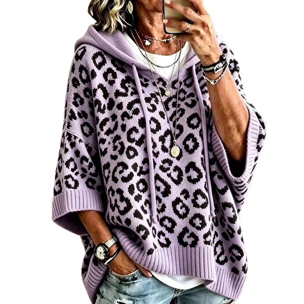 A woman wearing a lavender purple and black leopard print knit hoodie sweater, paired with distressed blue jeans and layered necklaces, holding a phone to take a mirror selfie against a plain white background.