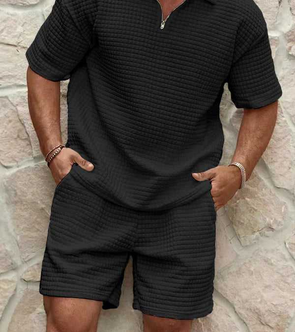 Person wearing a black textured outfit against a stone wall.