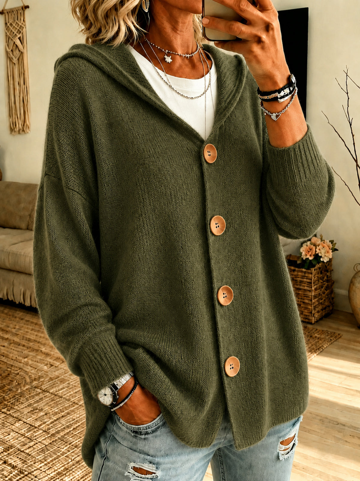 A woman wearing an olive green knit cardigan with a hood and large brown buttons, layered over a white top and paired with distressed light blue jeans in a cozy indoor setting.