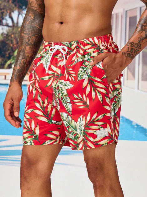 Person wearing red tropical leaf patterned swim shorts by a pool.