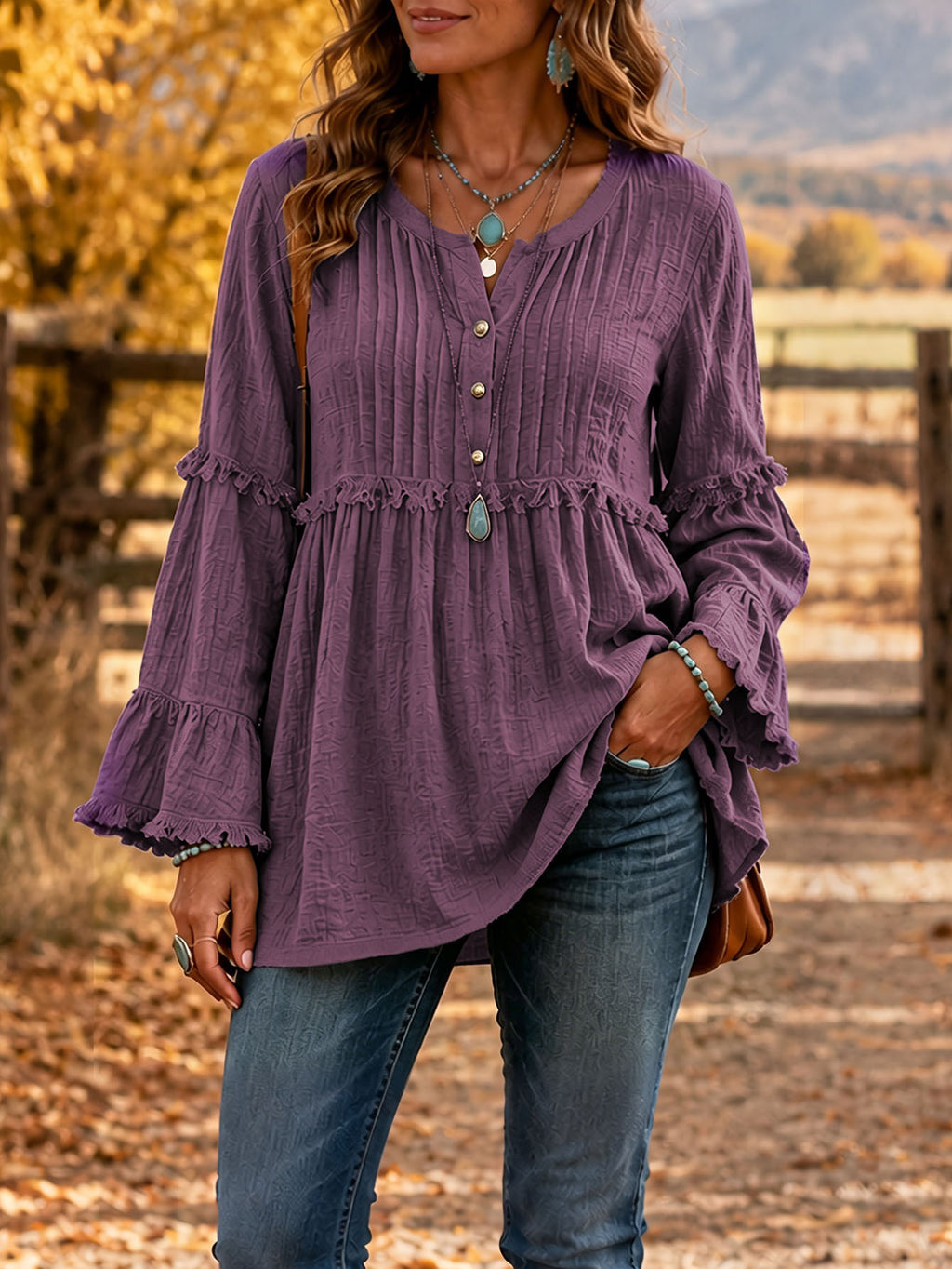 A woman wearing a purple long-sleeve blouse with a buttoned front and ruffled cuffs, paired with blue jeans, standing outdoors in a rustic autumn setting.