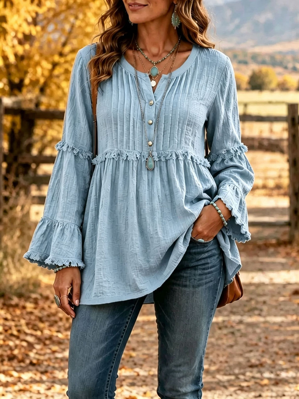 A woman wearing a light blue long-sleeve blouse with a gathered peplum waist, ruffled cuffs, and front buttons, paired with blue jeans in an outdoor autumn setting.
