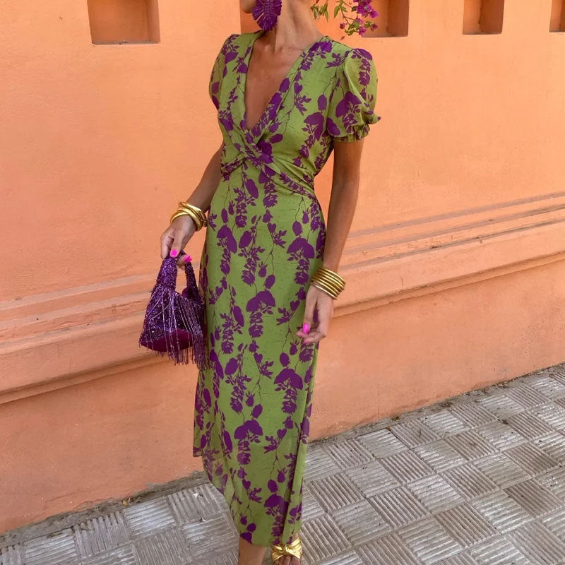 Woman in a green and purple floral dress holding a matching handbag against an orange wall.