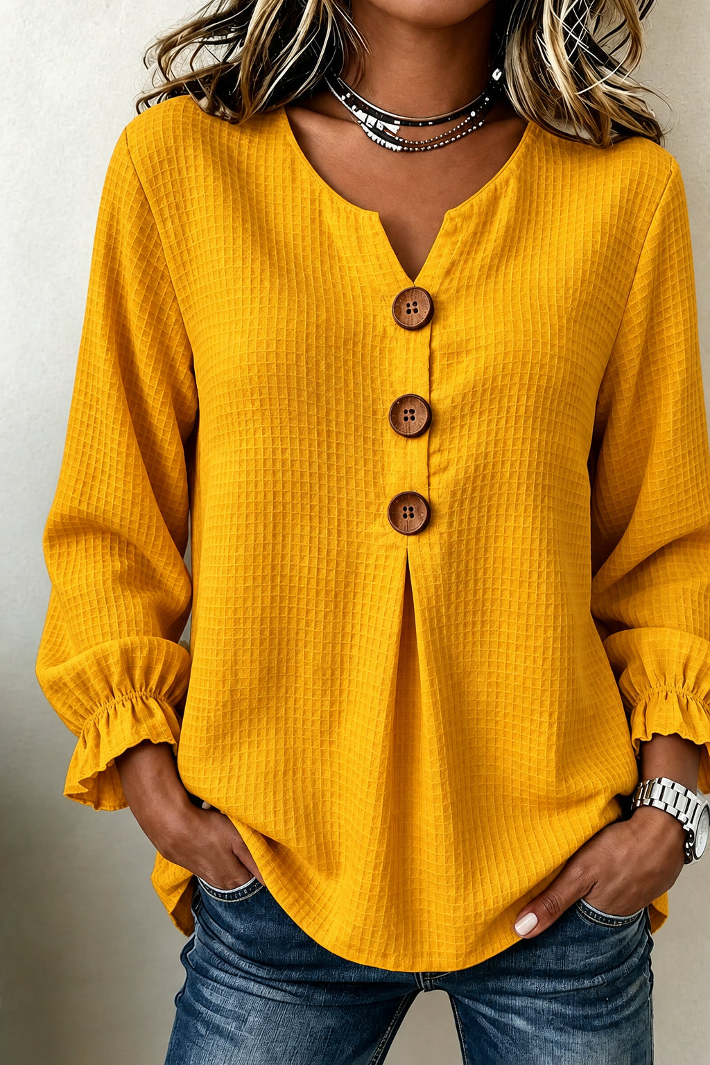 A woman wearing a mustard yellow waffle-knit long-sleeve blouse with a split V-neck, three brown buttons, and ruffled cuffs, paired with blue jeans.