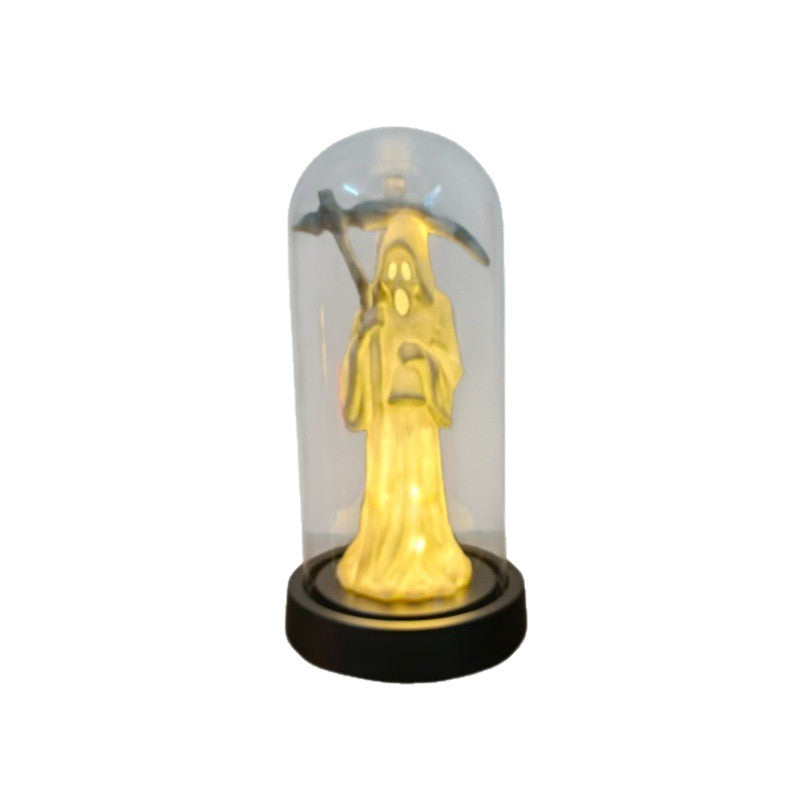A small decorative figurine of a hooded ghostly figure holding a pickaxe, illuminated from within in a warm yellow light, enclosed in a clear glass dome on a round black base.