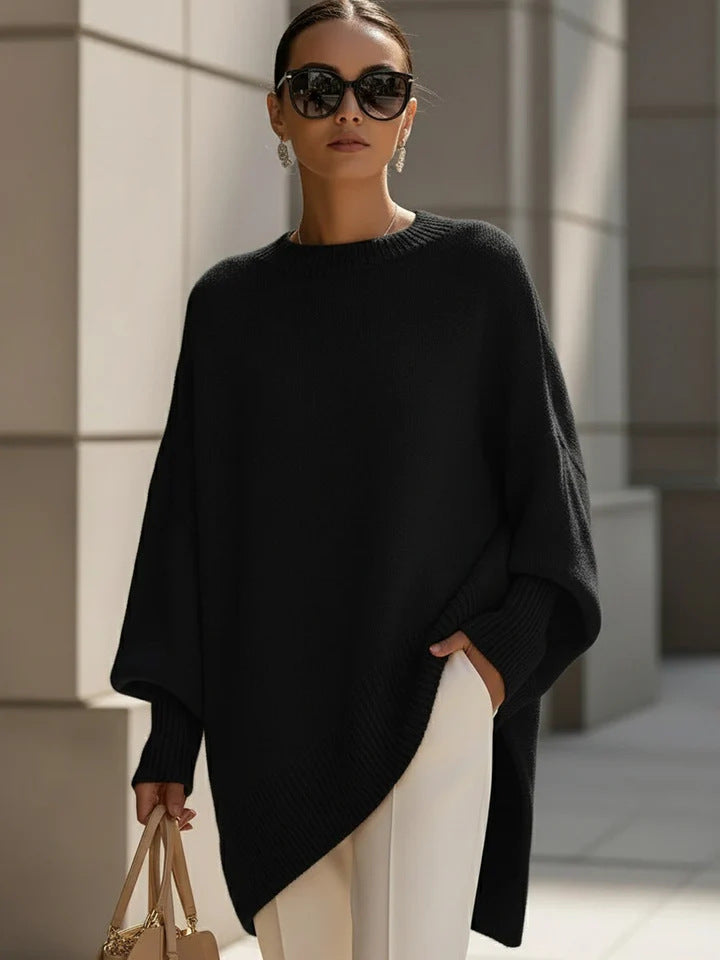 Woman wearing a black poncho and sunglasses, standing against a light-colored wall.