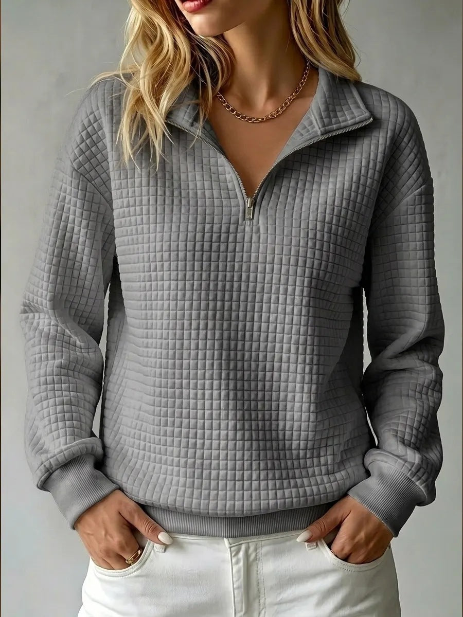 A woman wearing a gray waffle-textured quarter-zip pullover sweater with a stand collar, paired with white pants, shown against a neutral gray background.