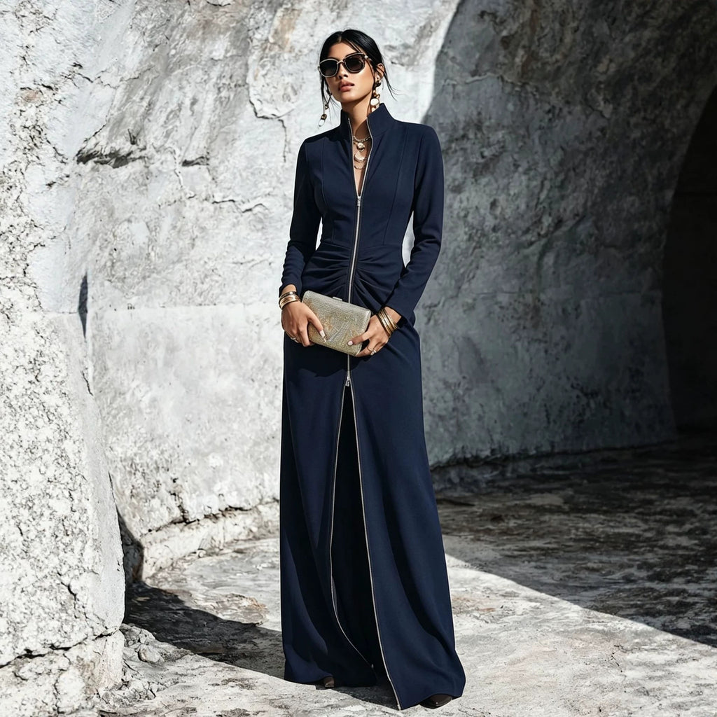 A woman wearing a long navy maxi dress with a front zipper, standing in a rocky stone corridor and holding a metallic clutch.