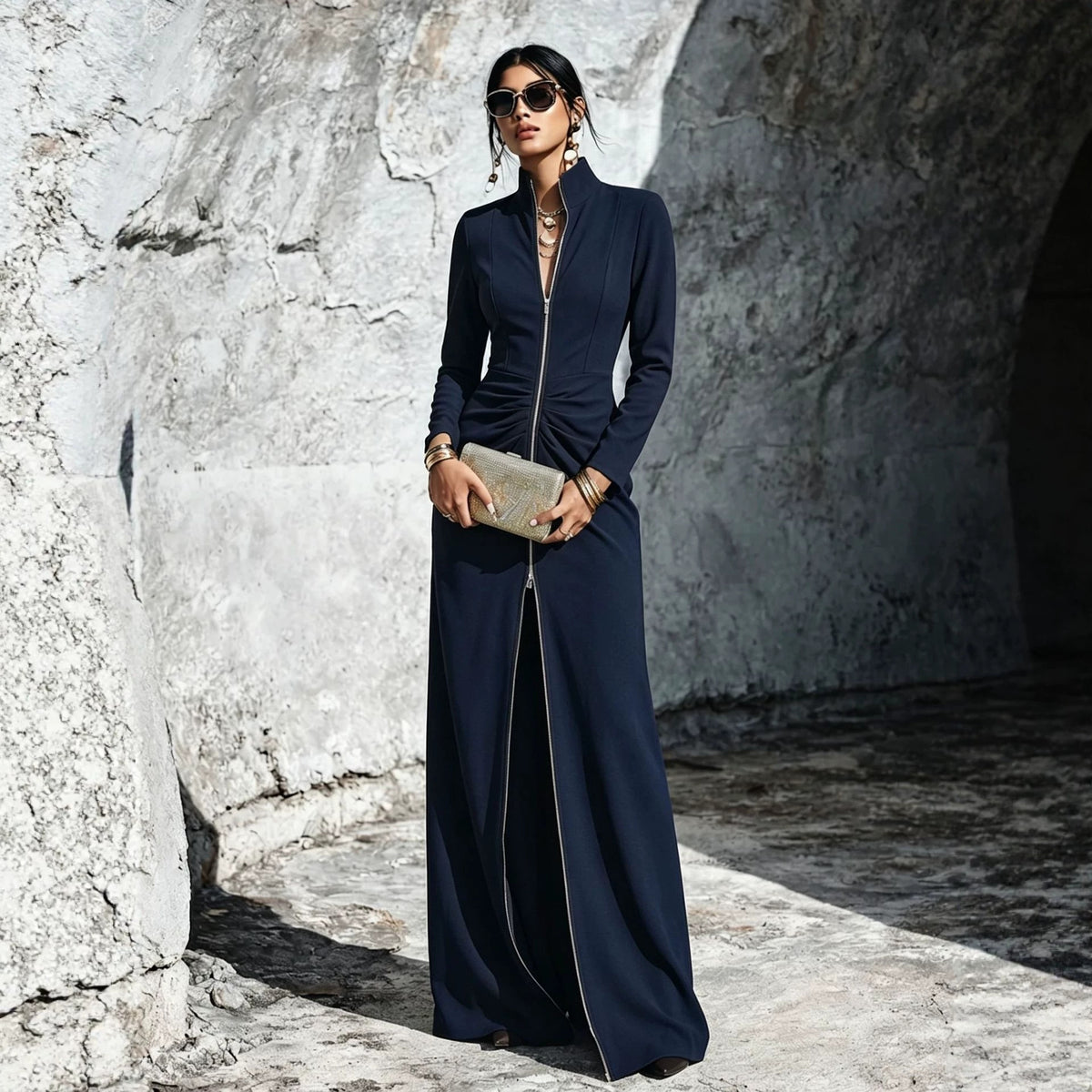 A woman wearing a long navy maxi dress with a front zipper, standing in a rocky stone corridor and holding a metallic clutch.