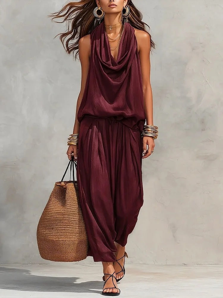 Woman wearing a burgundy sleeveless top and harem pants, holding a woven bag against a neutral background.