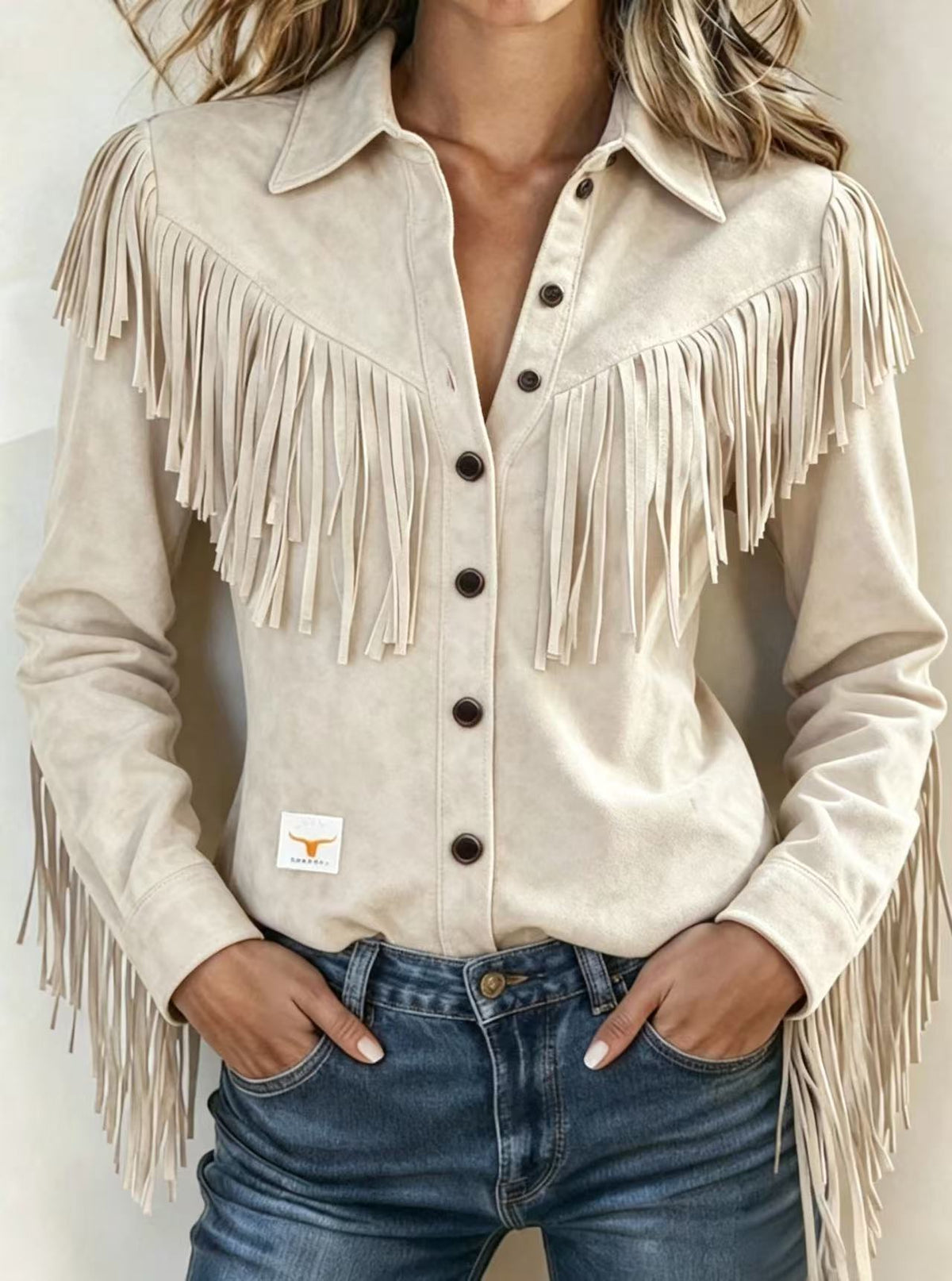 A woman wearing a beige suede-like long-sleeve button-up shirt with large fringe across the chest and shoulders, paired with blue jeans.
