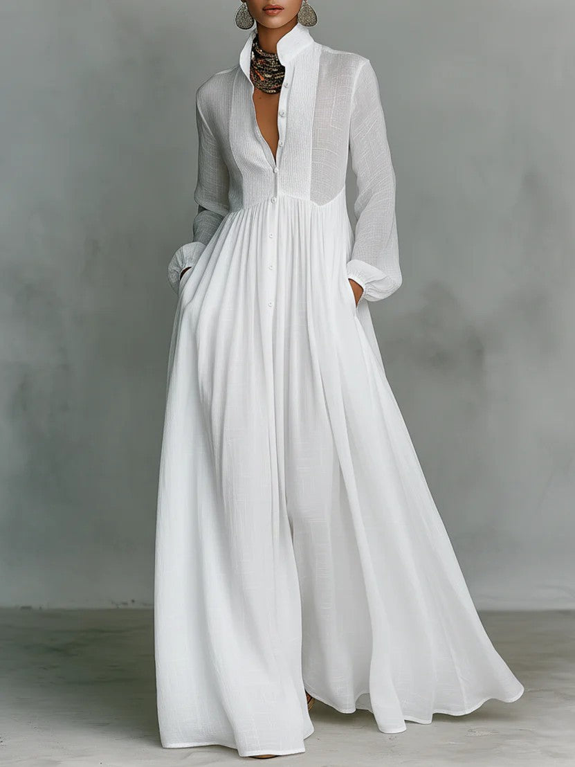 A woman models a long-sleeve white maxi dress with a deep V-neckline, sheer yoke, and button-front waist, set against a neutral gray studio backdrop.