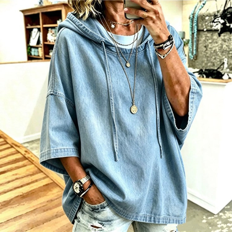 A person takes a mirror selfie wearing an oversized light blue denim hooded top with drawstrings, layered necklaces, bracelets, and distressed light-wash jeans in an indoor boutique setting.