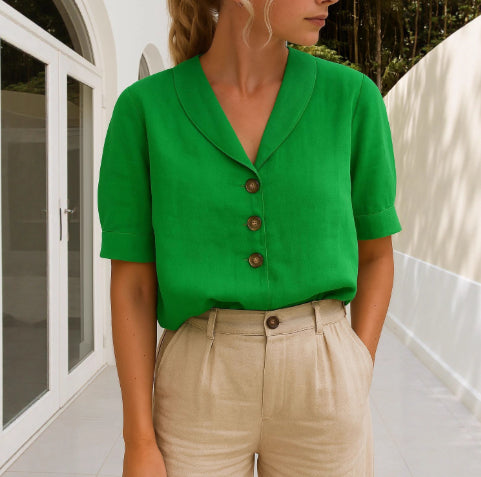 A woman wearing a bright green short-sleeve button-front top with a wide collar and beige high-waisted trousers, standing in a sunlit outdoor corridor.