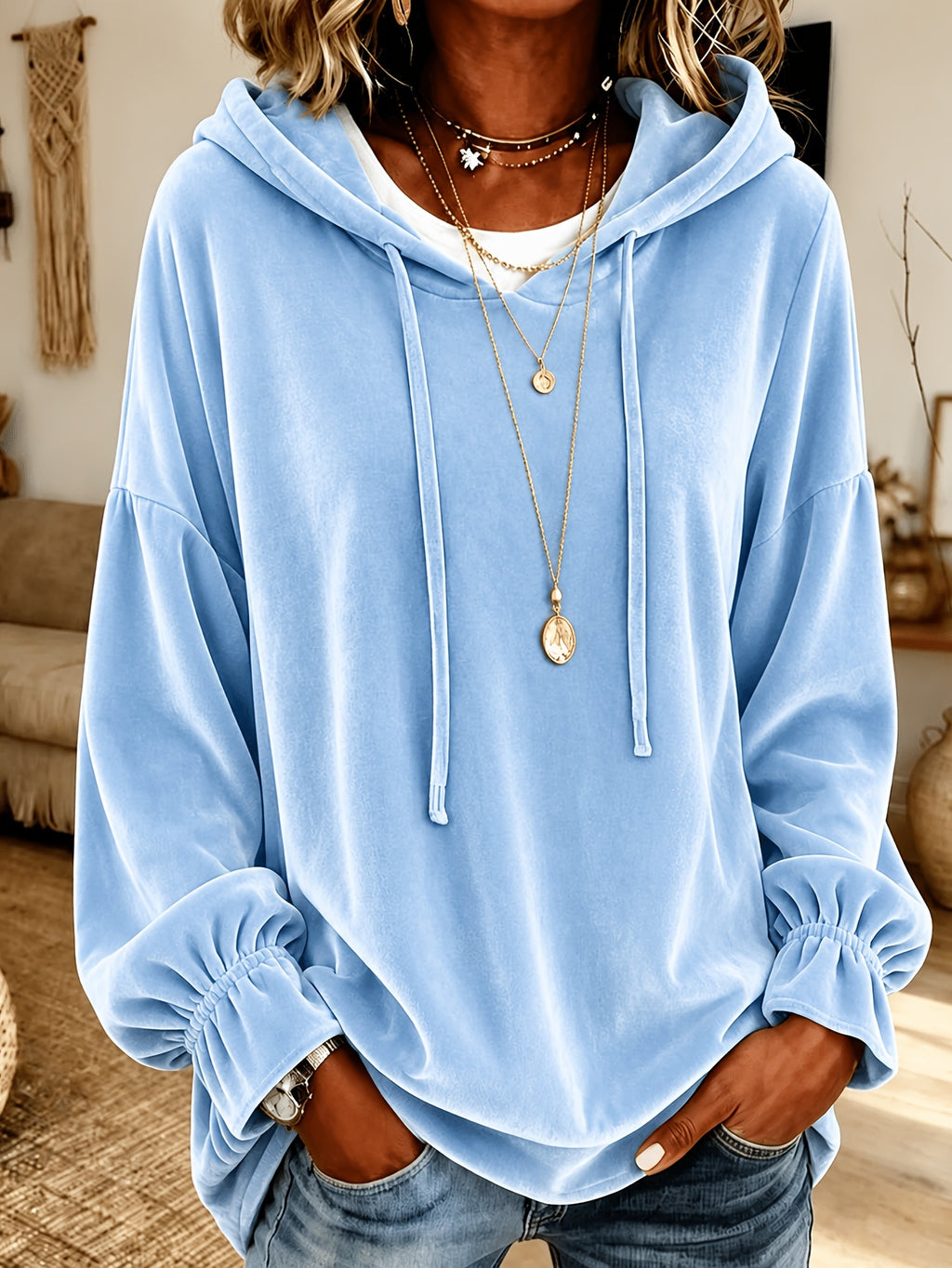 A person wearing a light blue velour hoodie with drawstrings and gathered cuffs, layered over a white undershirt and accessorized with layered gold necklaces, paired with blue jeans in a warm indoor setting.
