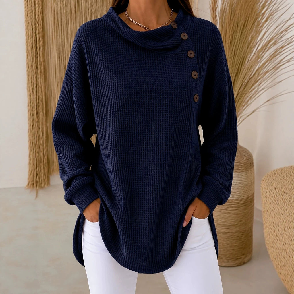 A woman wearing a navy blue waffle-knit cowl-neck sweater with an asymmetrical button closure, paired with white pants, standing in a styled indoor setting with woven decor.