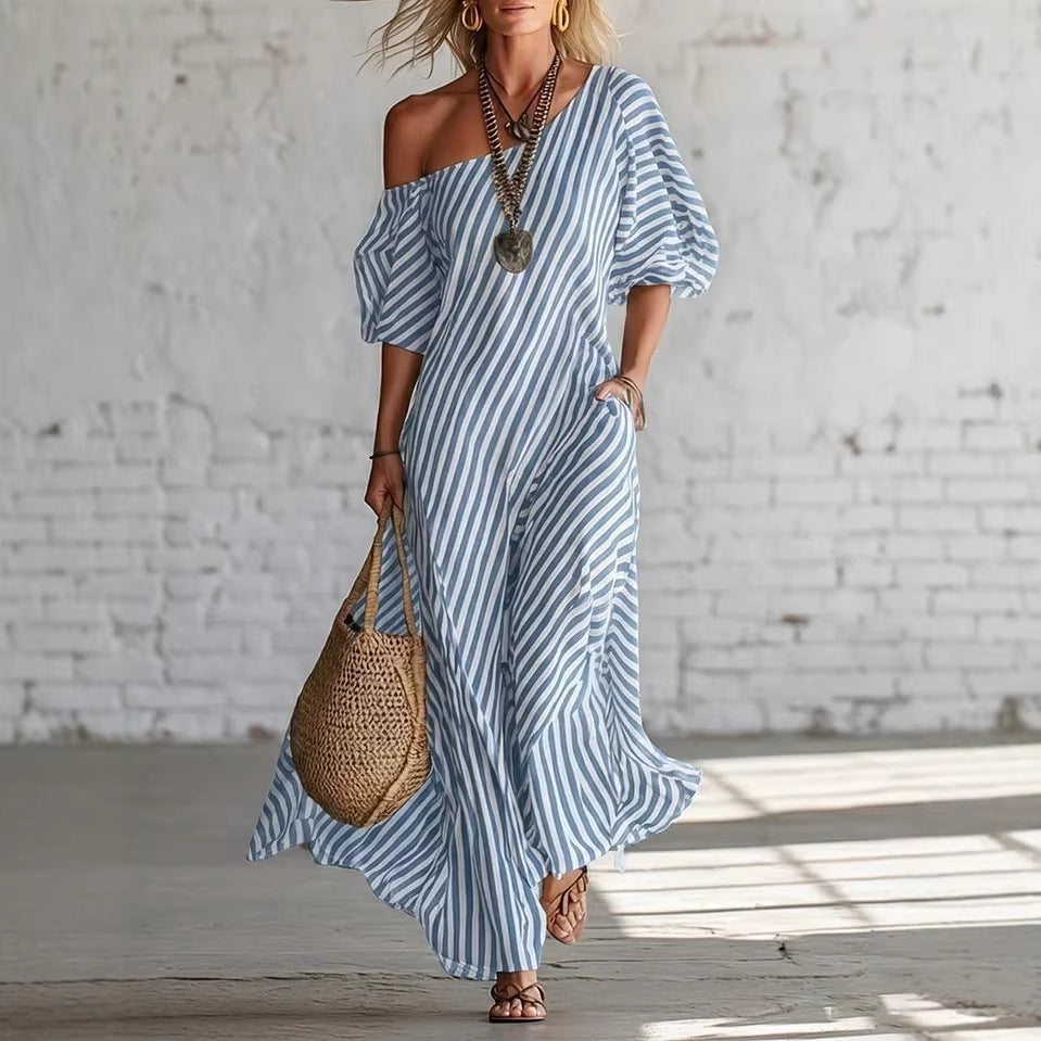 Woman wearing a blue and white striped dress holding a woven bag against a white brick wall.