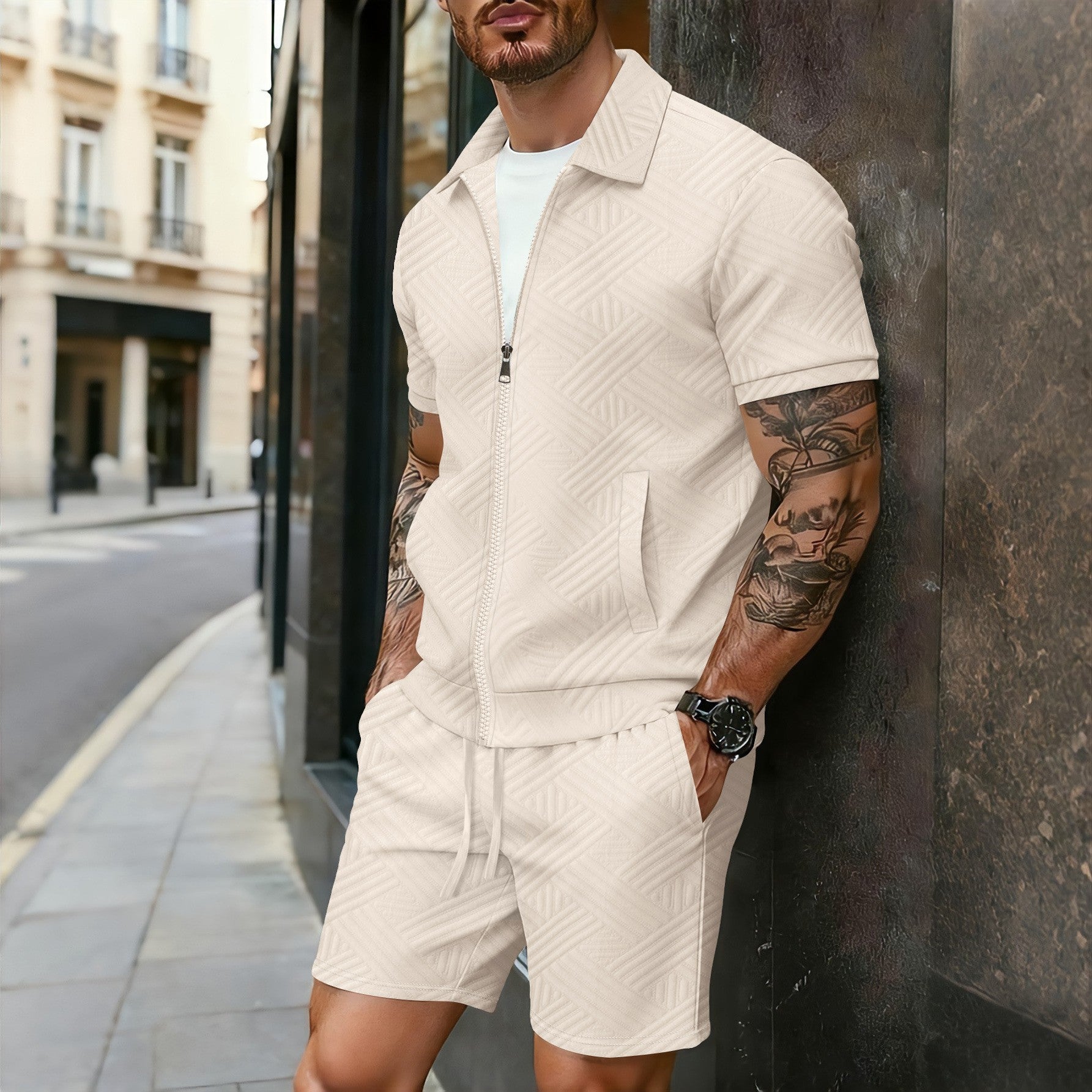 Man wearing a beige quilted jacket and shorts leaning against a wall in an urban setting.