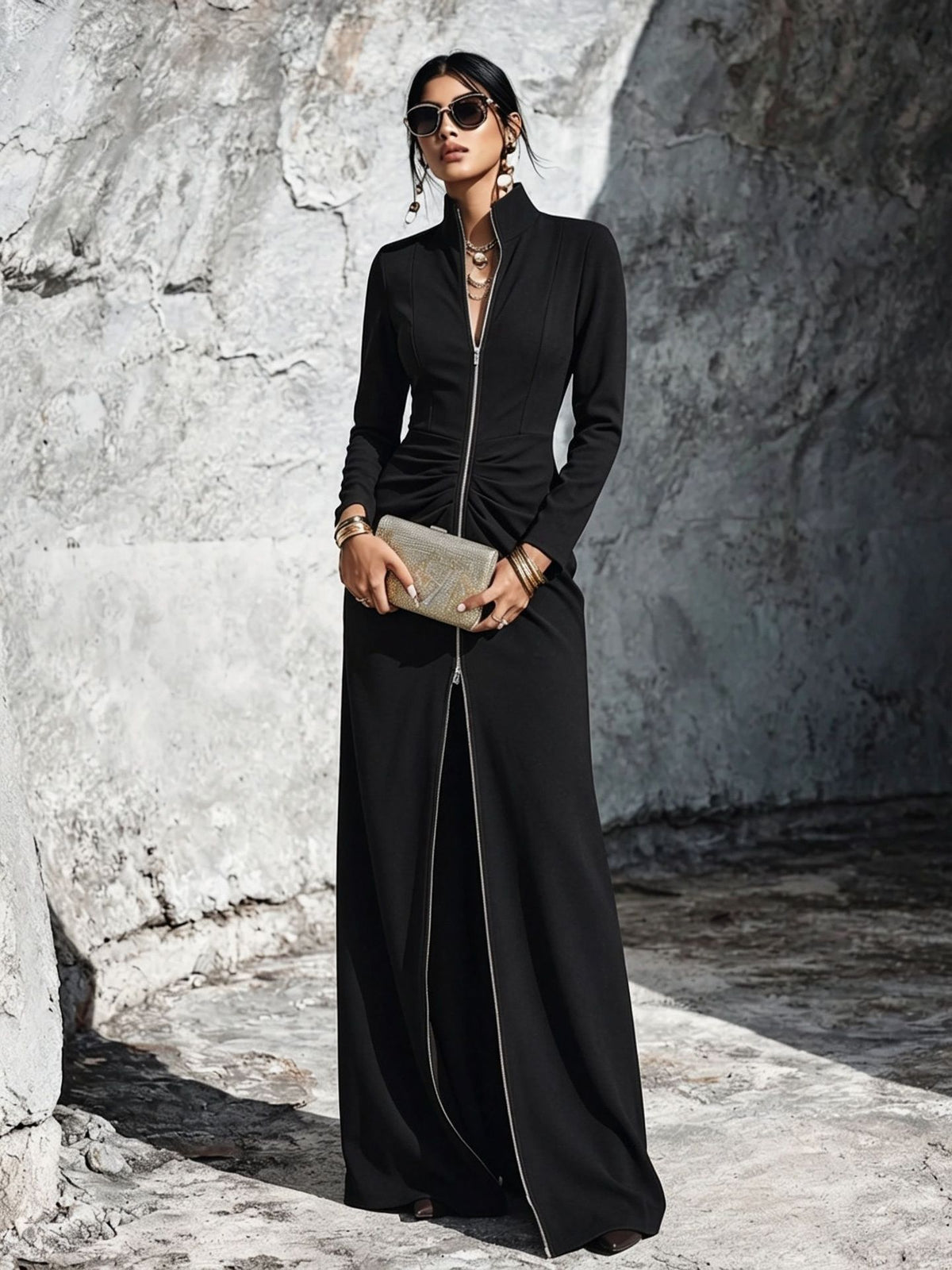 A woman wearing a long black maxi dress with a front zipper and gathered waist, styled with gold jewelry and a metallic clutch, standing against a textured gray stone wall.