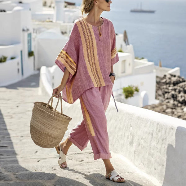 Woman in a pink outfit with yellow stripes walking on a sunlit street with white buildings and blue sea in the background.