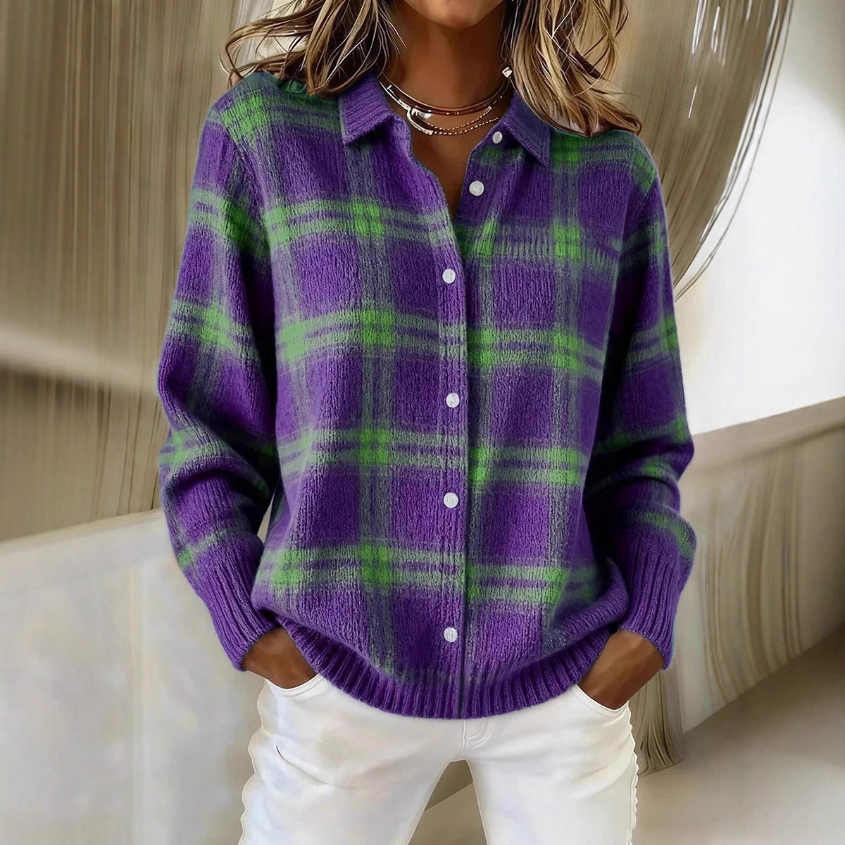 A woman wearing a purple and green plaid knit button-up sweater with white pants, standing in an indoor setting with a modern curved wall in the background.