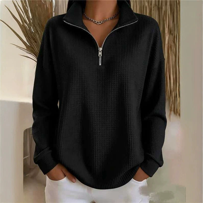 A person wearing a black quarter-zip long-sleeve top with a textured waffle knit pattern and white pants, standing indoors against a light neutral wall with dried plant decor.