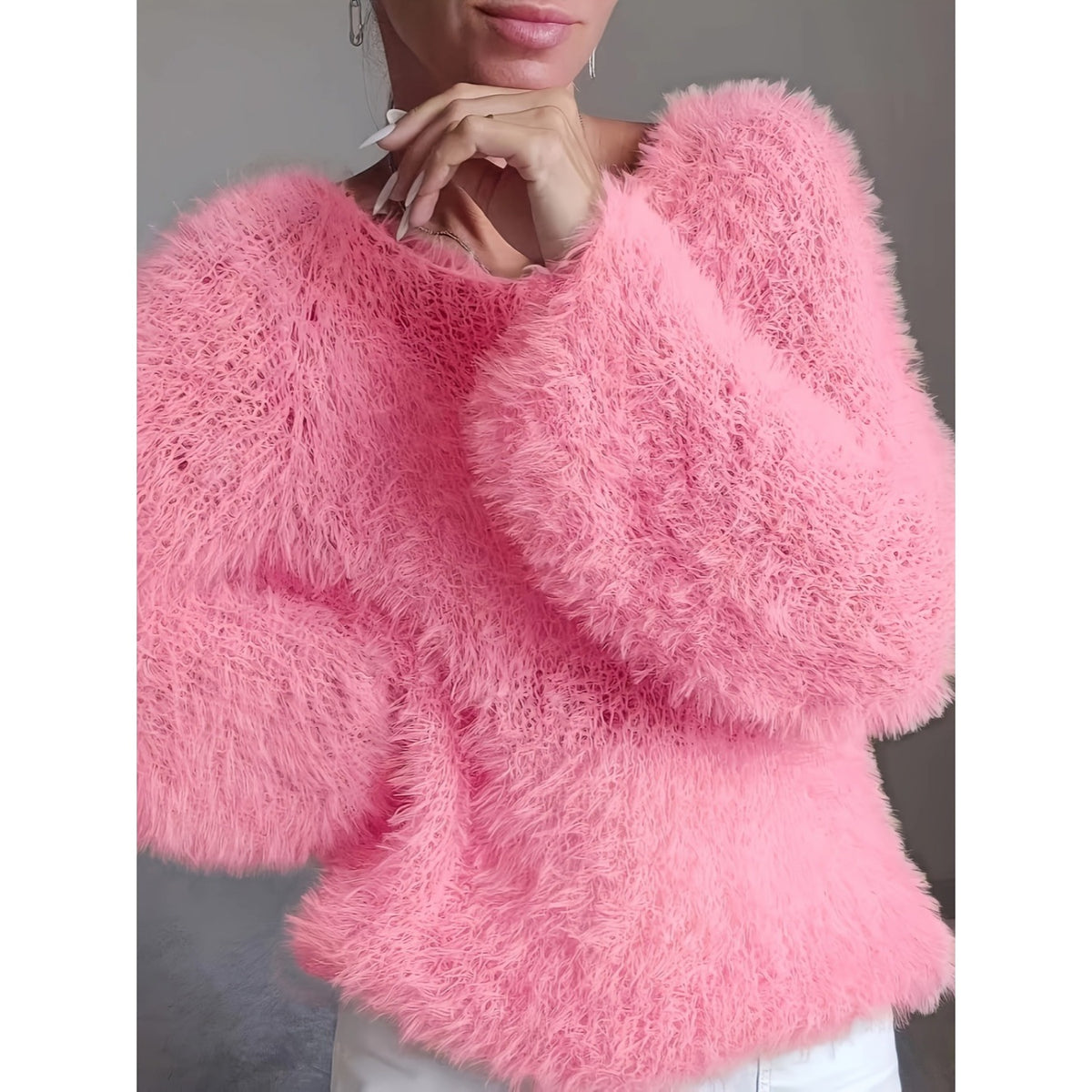 A close-up of a person wearing a bright pink, long-pile fuzzy knit sweater with voluminous balloon sleeves, paired with white pants against a neutral gray background.
