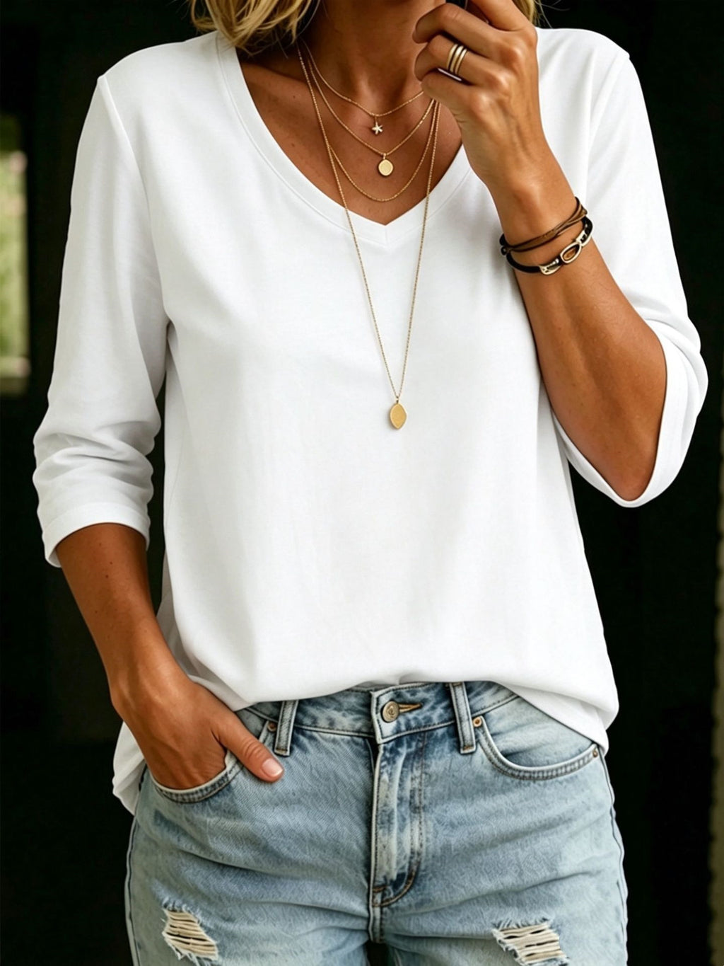 A woman wearing a white long-sleeve V-neck top with light blue distressed jeans, layered gold necklaces, and bracelets, shown from the neck down against a dark background.