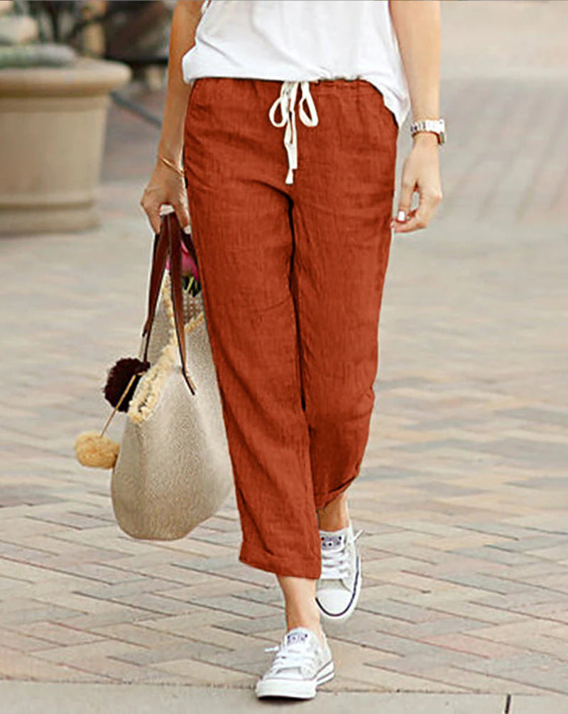 Person wearing rust-colored pants and white sneakers, holding a straw bag on a paved walkway.