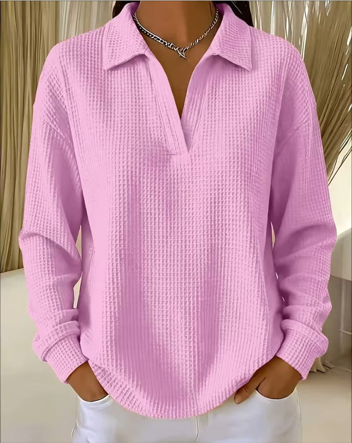 A woman wearing a light pink waffle-knit long-sleeve polo shirt with a split V-neck, paired with white pants and a silver chain necklace.