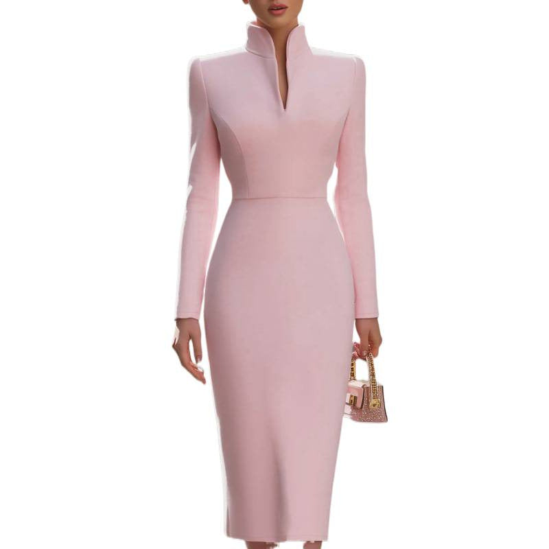 A woman modeling a fitted long-sleeve midi dress in light pink with a high mock neck and split neckline, holding a small matching pink handbag against a plain white background.