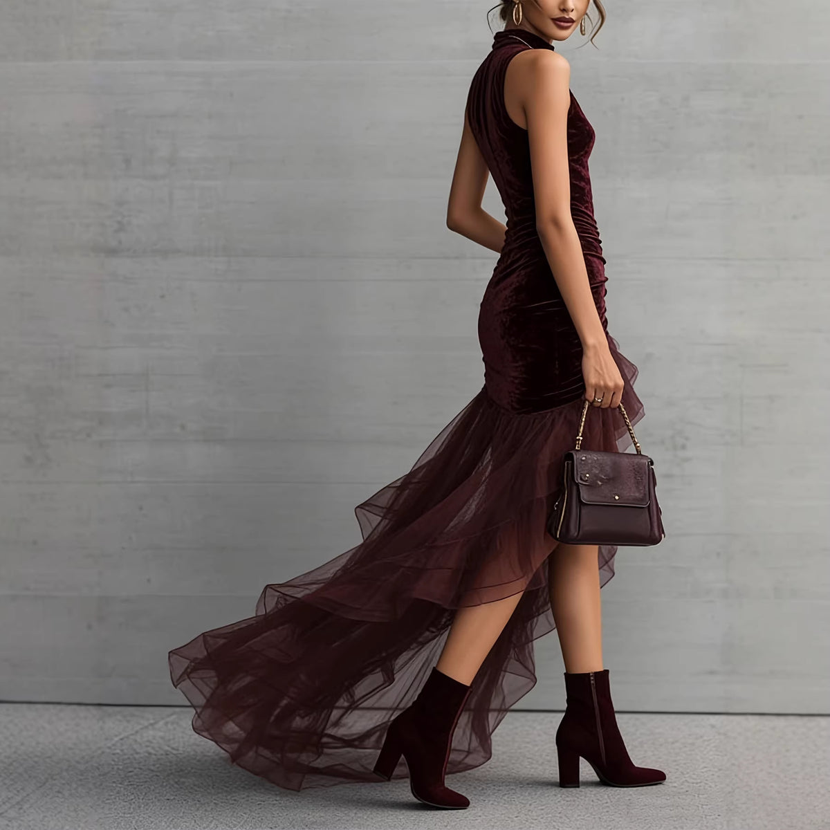 A woman models a sleeveless, high-neck, deep burgundy velvet dress with a tiered tulle skirt, paired with matching burgundy heeled ankle boots and a matching handbag, set against a gray concrete wall.