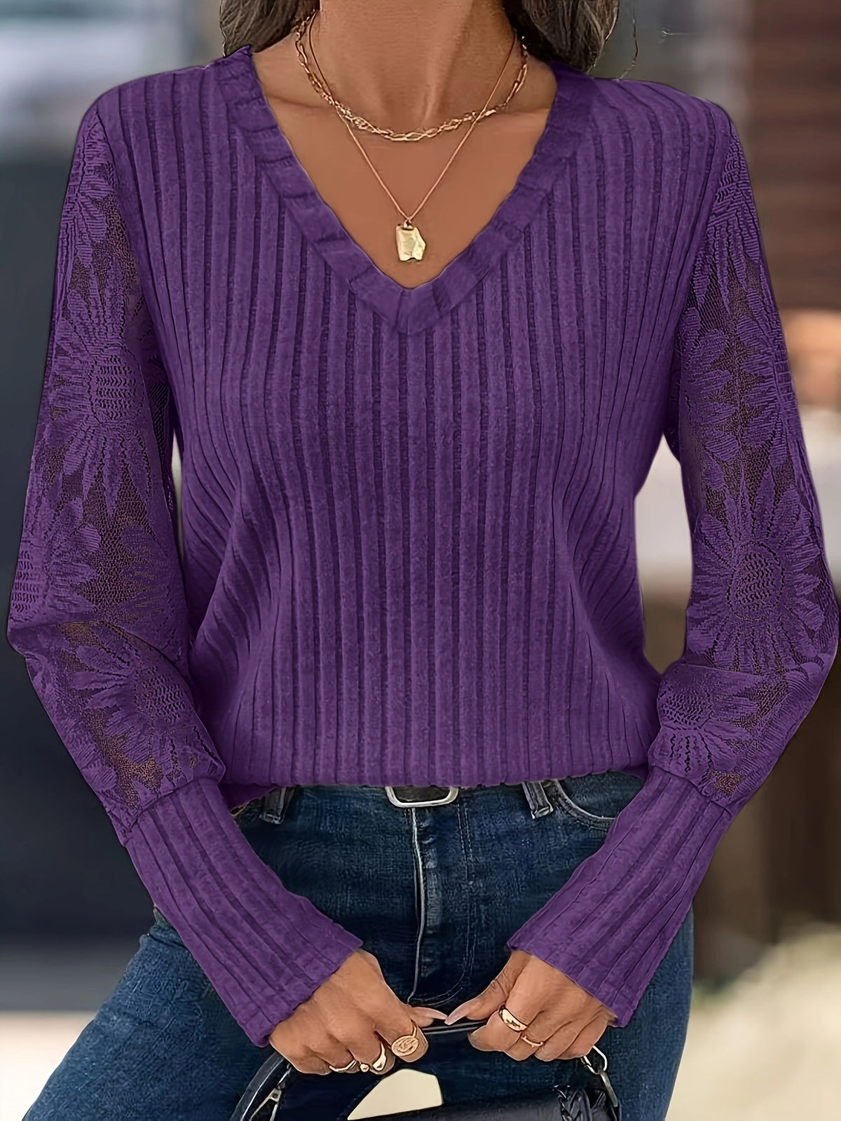 A woman wearing a purple ribbed knit sweater with lace sleeves and a V-neckline, paired with blue jeans and layered gold necklaces.